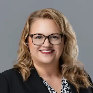 Studio headshot of a blonde woman in glasses and a black blazer on a gray backdrop