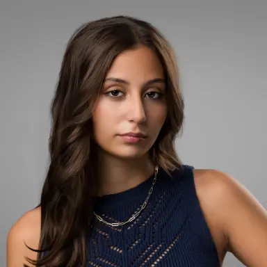 Studio headshot of a young woman with long dark hair in a navy sleeveless top on a gray backdrop