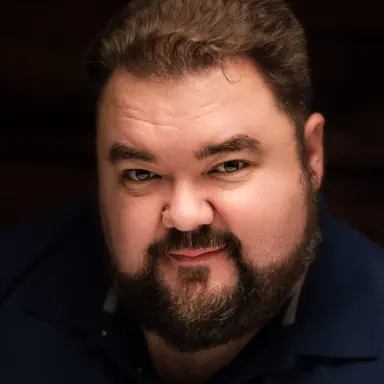 Studio headshot of a bearded man in a dark shirt on a dramatic dark backdrop