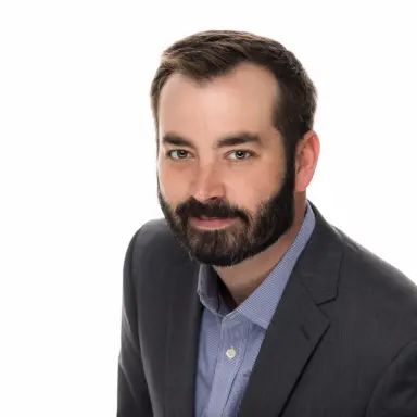Studio headshot of a bearded man in a gray blazer over a blue shirt on a white backdrop