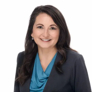 Studio headshot of a woman with dark hair in a navy blazer over a teal top on a white backdrop