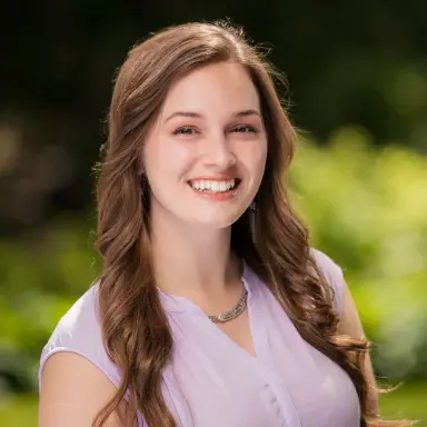 Outdoor headshot of a young woman with long brown hair in a lavender top with a green background