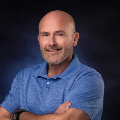 Studio headshot of a man with a shaved head and beard in a blue polo on a blue backdrop